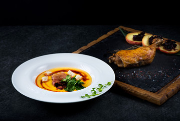 An appetizing pumpkin soup is served in a white plate in a restaurant. Duck confit with baked apple