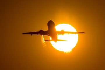 Silhouette of airplane taking off during sunset, flies through the disc of the sun. A trail of hot air from engines is visible.