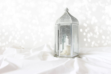 Ornamental silver Moroccan, Arabic lantern on white linen throw. Burning candle, glittering bokeh lights. Greeting card for Muslim community holy month Ramadan Kareem. Festive background.