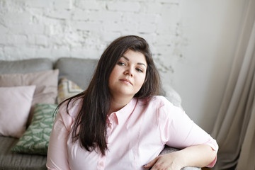 Naklejka premium Close up of attractive chubby girl in her twenties resting at home after she finished doing housework, sitting on couch in living room, thinking about something. People, rest, leisure and relaxation