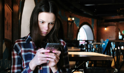 Upset woman sends a message or use internet in the phone sitting in a cafe. Dressed in a plaid shirt.