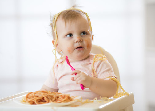 Funny child eating noodle. Grimy kid girl eats spaghetti with fork sitting on table at home