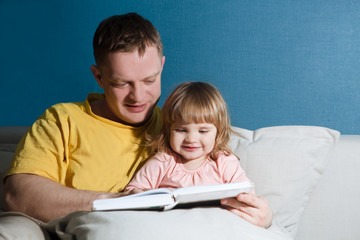 Father reading a story to his child girl sitting on couch at home. Happy family time together at home.
