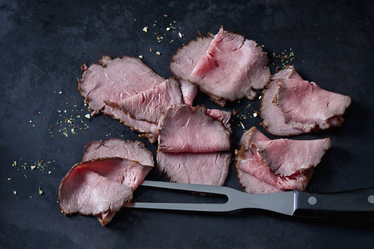 Six slices of roast beef, herbs and meat fork on dark ground