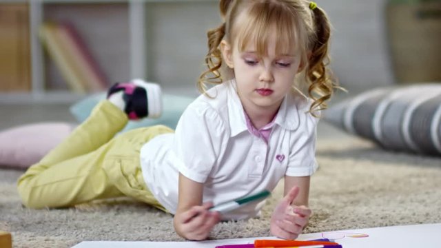 Adorable little girl with wavy golden hair and two ponytails lying on the floor and using colored felt-tip pens to draw a picture
