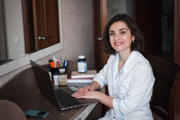 Obraz na płótnie Canvas A young smiling brunette doctor woman in white medical gownsits at her desk, which has a laptop, books, accessories and jars of medicines or vitamins.