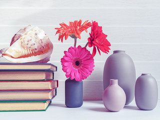 The seashell is on a stack of books. Three gerbera flowers are in a gray vase. Bottles of different shapes and sizes are near. Light wooden background