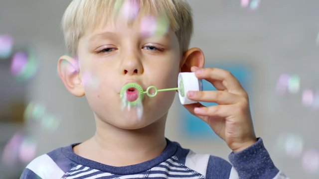 Close up face of cute primary school boy blowing soap bubbles at camera with bubble blower toy