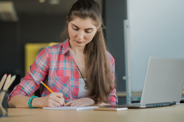 Portrait of a Serious Woman Writing in the Notebook while Working on Laptop lndoors.