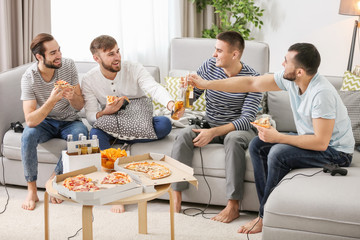 Young men drinking beer and eating pizza while playing video games at home