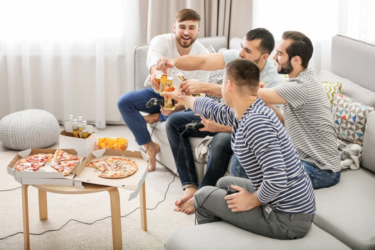 Young men drinking beer and eating pizza while playing video games at home