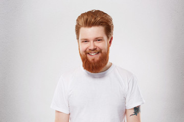 Fototapeta premium Young redhead man with heavy, massive beard smiles and feels happy. Portrait of pleased, joyful male. Isolated over studio white wall