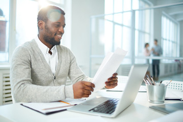 Waist-up portrait of cheerful African American businessman studying document while sitting at desk...