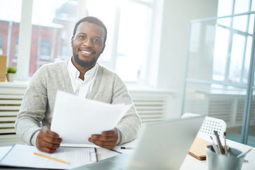 Waist-up portrait of smiling African American manager looking at camera while sitting at office desk and finishing promising project, interior of modern office on background