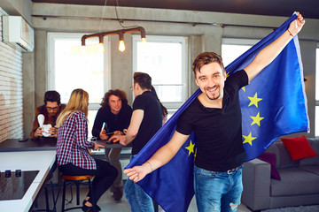 Young man with the european flag in his hands on the background of friends in the room.