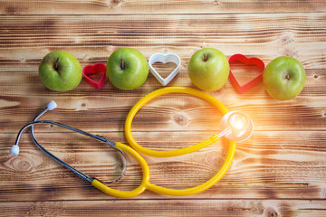 The green apple with heart plastic model stack on wooden timber board,at the back of yellow stethoscope,vintage warm light tone,blurry light around,health care concept