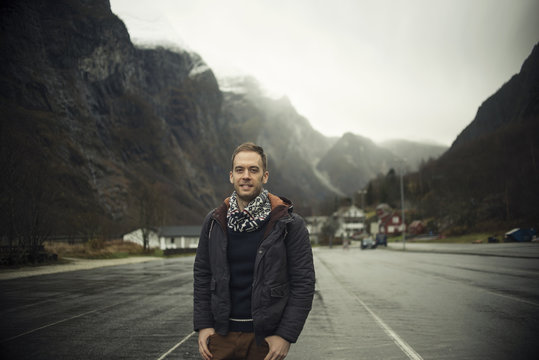Retrato de hombre blanco joven rubio y chaquetón azul en una carretera frente a unas montañas y un pequeño pueblo