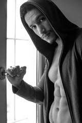 sexy and naked muscular young man in jacket with hood standing near the window