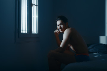 sexy and naked muscular young man sits on a bed in the room. the light from the window
