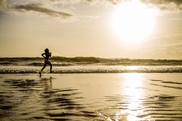 silhouette of young Asian sport runner woman in running workout training at sunset beach with orange sunlight reflection on the sea water