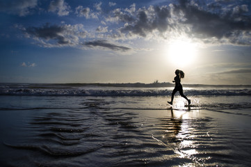Fototapeta premium silhouette of young Asian sport runner woman in running workout training at sunset beach with orange sunlight reflection on the sea water
