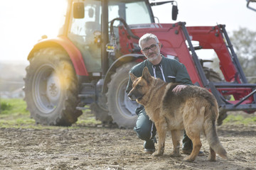Farmer petting dog outside the barn, tractor in background