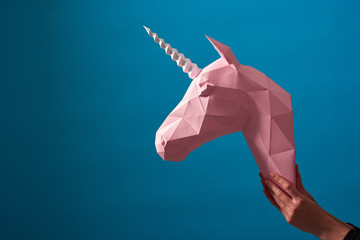 Pink paper unicorn's head in woman's hands with accurate manicure on blue background. Intresting geometrical lines and shape. Original interior accessory. Modern decor detail.