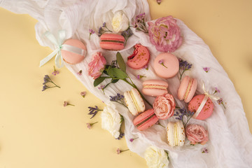 Pink and white macaroons cakes with big and small flower buds are decoratively laid on a white fabric on a yellow background