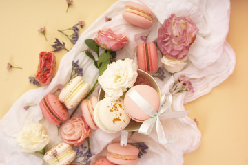 Obraz na płótnie Canvas Pink and white macaroons cakes with big and small flower buds are decoratively laid on a white fabric and a cup on a yellow background