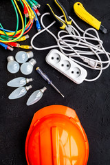 Electrician work concept. Hard hat, tools, cabel, bulb, socket outlet on black background top view