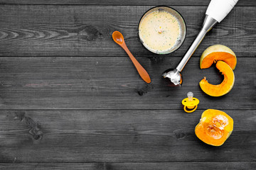 Make baby food at home. Puree with pumpkin near immersion blender and pacifier on dark wooden background top view copy space
