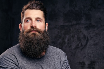 Portrait of a hipster with full beard and stylish haircut, dressed in a gray t-shirt, stands with a thinking look in a studio on a dark background.