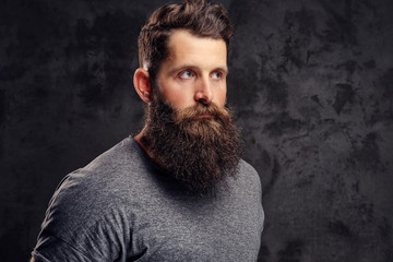 Portrait of a hipster with full beard and stylish haircut, dressed in a gray t-shirt, stands with a thinking look in a studio on a dark background.