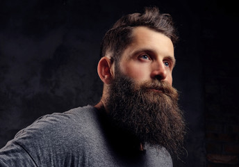 Portrait of a hipster with full beard and stylish haircut, dressed in a gray t-shirt, stands with a thinking look in a studio on a dark background.