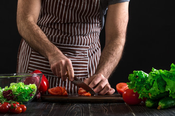 Man preparing salad on a wooden table. Men's hands cut the tomato to make a salad on black background