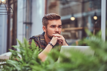 Portrait of a smiling fashionable bearded male with a stylish haircut, dressed in a flannel shirt, sitting in a cafe outdoors.