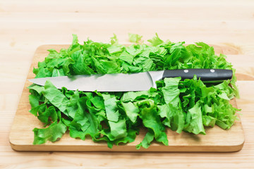 Close-up on a wooden table is a wooden cutting board on which lies the chopped lettuce leaves and on them lies a large cutting knife. The concept of self-cooking vegetarian food at home. Healthy