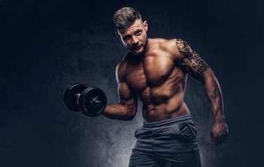 Naklejka na ściany i meble A handsome shirtless tattooed bodybuilder with stylish haircut and beard, wearing sports shorts, posing in a studio. Isolated on a dark background