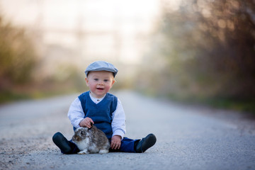 Adorable toddler boy, child playing with little bunny on a rural road with blooming trees
