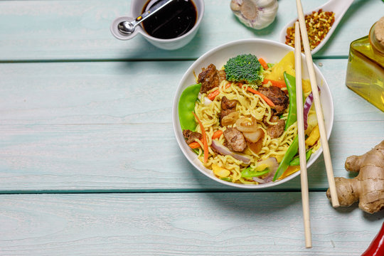 Chinese noodles with vegetables, mushrooms and meat in bowl on light blue wooden ruxtic table, top view