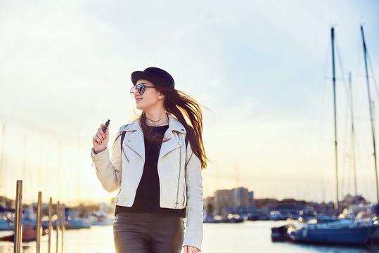 Pretty young woman in black hat with vape at a sea port