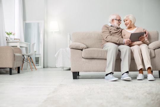 Modern Technology. Nice Joyful Elderly Couple Sitting Together On The Sofa While Using A Tablet