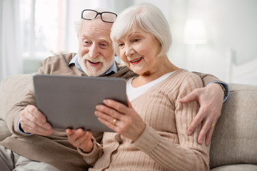 Modern tablet. Nice pleasant senior couple sitting together on the sofa while using modern tablet - obrazy, fototapety, plakaty