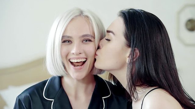 Young girl is kissing in cheek her female friend unexpected in a bedroom. Blonde is wondering and enjoying, opening mouth and showing tongue, close-up