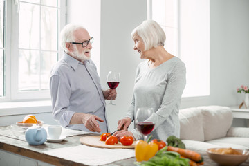 Delicious dinner. Positive senior man holding a glass of wine while talking to his wife