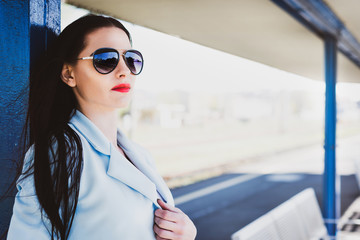 Portrait of a beautiful attractive woman with red lips. Cute girl in a blue jacket and a concentrated pose posing on the platform of a railway station. Female in sunglasses.