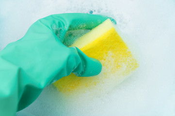 A hand in a turquoise glove holds a sponge for washing dishes in a copious foam. Close-up