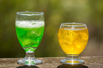two glasses with exotic green and yellow carbonated drinks with ice on the boards, against the background of greenery