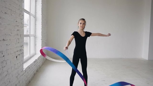 Attractive female gymnast dancing with ribbon in gymnastics school at white background and look at the camera. Rhythmic gymnast