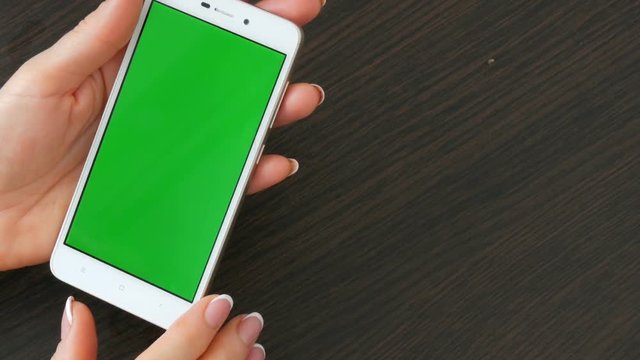 Female hands with beautiful French manicure take a white smartphone with Green Screen. Using Smartphone,Holding Smartphone with Green Screen on a stylish black wooden table